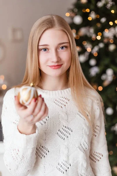 Mavi gözlü güzel sarışın kız elinde beyaz süveter giymiş bir Noel topunu Yeni Yıl ağacının arka planında tutuyor. Yılbaşı fotoğrafları. Noel fotoğrafı — Stok fotoğraf