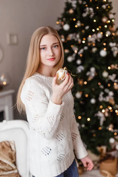 Menina loira bonita com olhos azuis segura em suas mãos uma bola de Natal em uma camisola branca contra o fundo da árvore de Ano Novo. Fotos de Ano Novo. Foto de Natal — Fotografia de Stock