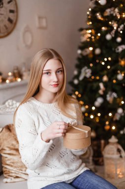 Mavi gözlü güzel sarışın kız yeni yıl ağacının arka planında beyaz bir süveter içinde elinde bir hediye tutuyor. Yeni yıl fotoğrafları
