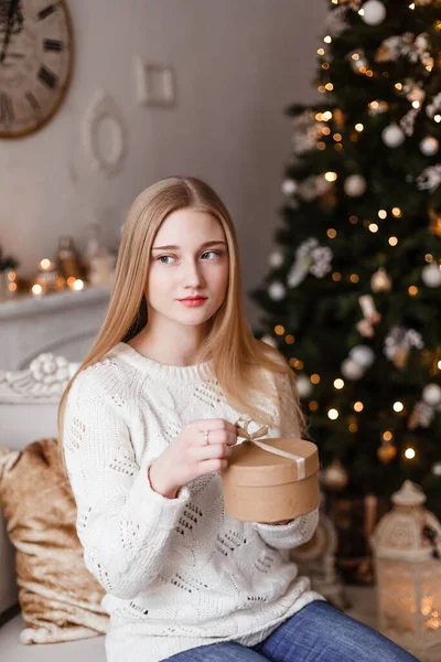 Schöne blonde Mädchen mit blauen Augen hält ein Geschenk in ihren Händen in einem weißen Pullover auf dem Hintergrund des Neujahrsbaums. Neujahrsfotos — Stockfoto