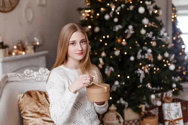 Piękna blondynka z niebieskimi oczami trzyma w dłoniach prezent w białym swetrze na tle drzewa noworocznego. Zdjęcia noworoczne — Zdjęcie stockowe