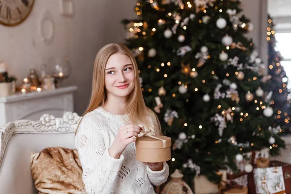 Piękna blondynka z niebieskimi oczami trzyma w dłoniach prezent w białym swetrze na tle drzewa noworocznego. Zdjęcia noworoczne — Zdjęcie stockowe