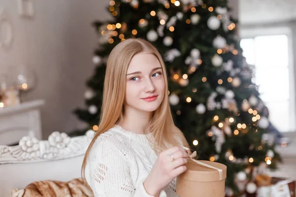 파란 눈을 가진 아름다운 금발의 소녀 가새년 나무의 배경에 하얀 스웨터를 입고 손에 선물을 들고 있다. New Years photos — 스톡 사진