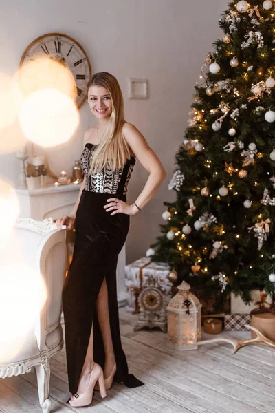 Una rubia lujosa en un vestido largo de terciopelo negro se para en el árbol de Navidad decorado y sonríe, preparándose para celebrar el Año Nuevo y la Navidad. —  Fotos de Stock