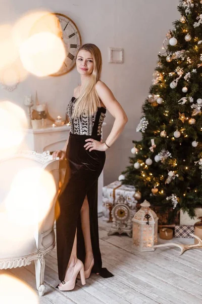 Una lussuosa bionda in un abito da sera di velluto nero si erge sull'albero di Natale decorato e sorridente, preparandosi a festeggiare il nuovo anno e il Natale. — Foto Stock