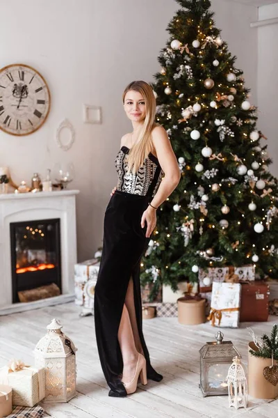 Luksusowa blondynka w czarnej aksamitnej sukni wieczorowej stoi na ozdobionej choince i uśmiecha się, przygotowując się do świętowania Nowego Roku i Bożego Narodzenia — Zdjęcie stockowe
