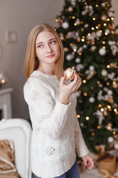 Piękna seksowna młoda kobieta w białym swetrze siedząca obok białej choinki, trzymająca prezent. Zdjęcie świąteczne — Zdjęcie stockowe