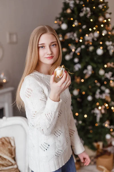 Schöne sexy junge Frau in weißem Pullover sitzt neben weißem Weihnachtsbaum und hält Geschenk. Weihnachtsfoto — Stockfoto