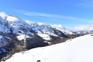 Güneşli bir günde karlı dağlarla kaplı kış manzarası. Karla kaplı bir yamaç ve dağ yolu manzaralı küçük bir köy. Ancare, Lugo, Galiçya, İspanya.