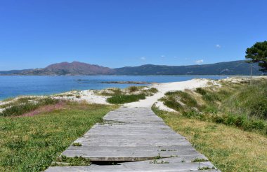 Tahta kaldırımlı sahil, kum tepeleri, turkuaz suyu ve mavi gökyüzü olan dağ ve koy. Carnota, İspanya.