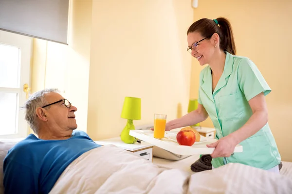Young smiling nurse brings breakfast to senior patient — Stock Photo, Image