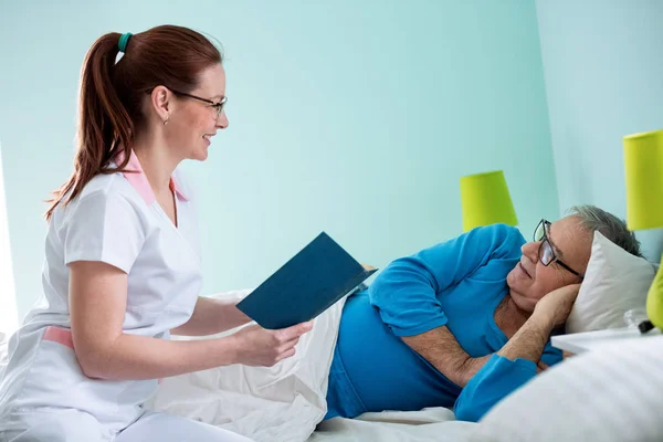 Smiling positive nurse reading a book to her old patient — Stock Photo, Image