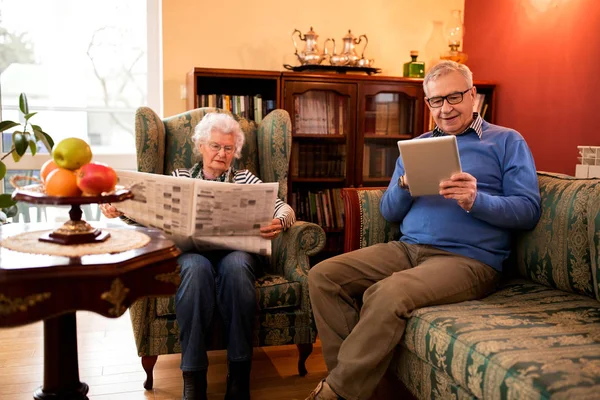 Senior couple relax at home while reading newspaper and using tablet