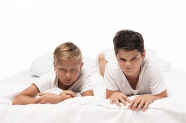 two angry boys grimacing while lying on bed and looking at camera isolated on white clipart