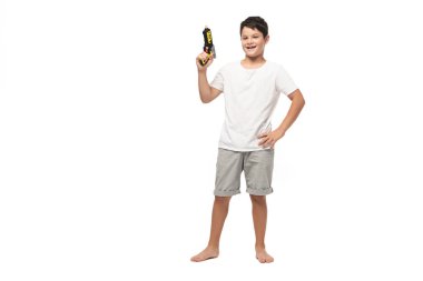 Elleri kalçasında duran ve beyaz arka planda kameraya gülümseyen oyuncak tabancayı tutan neşeli çocuk.