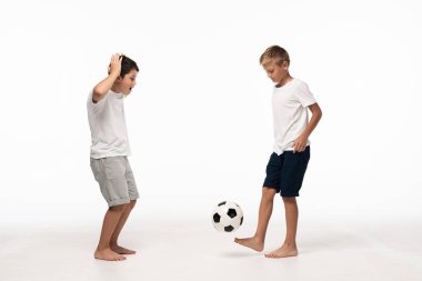 excited boy showing wow gesture while looking at brother playing with soccer ball on white background clipart