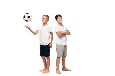 Gülümseyen kardeşin yanında futbol oynayan neşeli çocuk beyaz arka planda çapraz kollarla duruyor.
