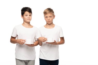 two sceptical brothers looking at camera while holding coffee cups on saucers isolated on white clipart