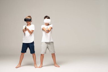 VR kulaklıklı iki kardeş, gri arka planda kameraya oyuncak silah doğrultuyor.