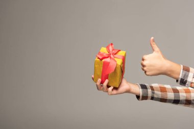 cropped view of boy holding yellow gift box and showing thumb up isolated on grey clipart