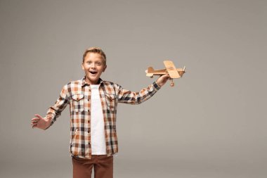 excited boy holding wooden toy plane and looking at camera isolated on grey clipart