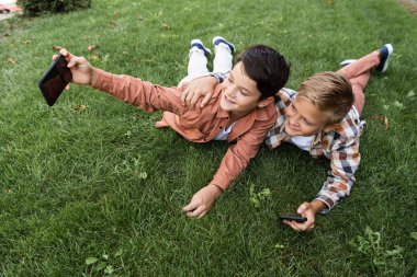 Neşeli çocuk, kardeşiyle çimde uzanırken akıllı telefondan selfie çekiyor.