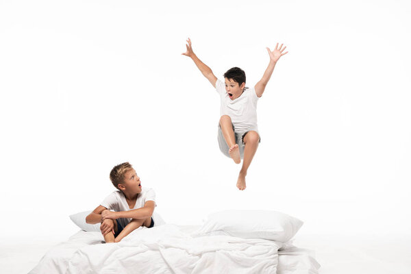 excited boy levitating over shocked brother sitting on bedding isolated on white