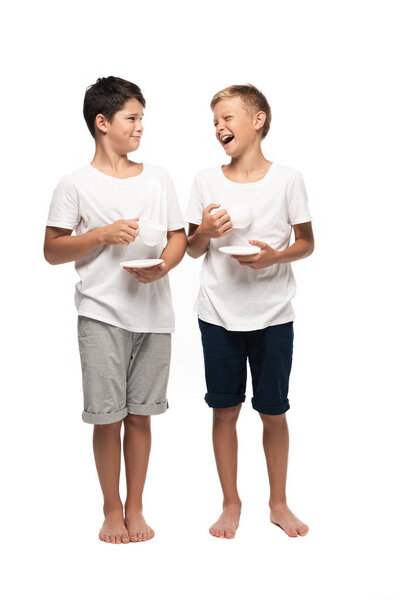 cheerful brothers looking at each other while holding coffee cups on white background