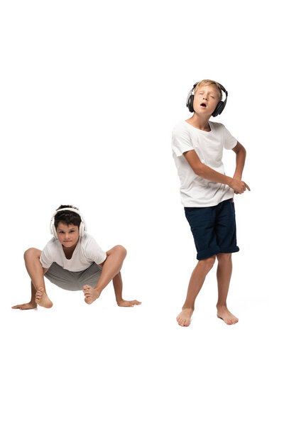 excited boy singing in headphones near brother sitting in frog pose on white background