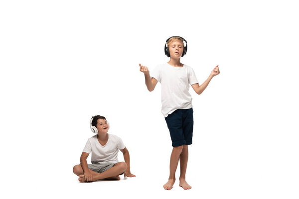 cheerful boy sitting with crossed legs and looking at brother dancing in headphones on white background