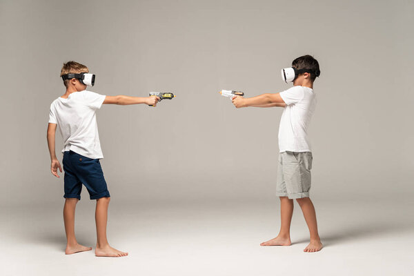 full length view of two brothers in vr headsets aiming at each other with toy guns on grey background