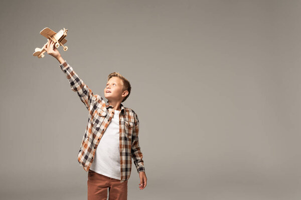 happy boy holding wooden toy plane in raised hand isolated on grey