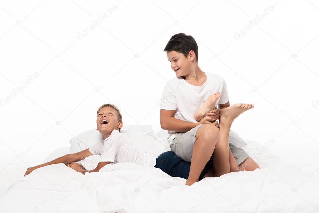 cheerful boy holding brothers leg while having fun on bed isolated on white
