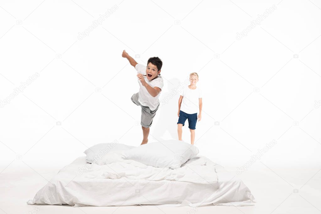 excited boy levitating over bed while smiling brother standing on white background