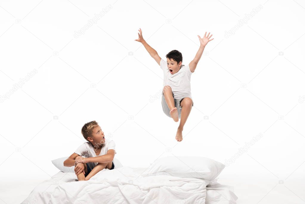 excited boy levitating over shocked brother sitting on bedding isolated on white