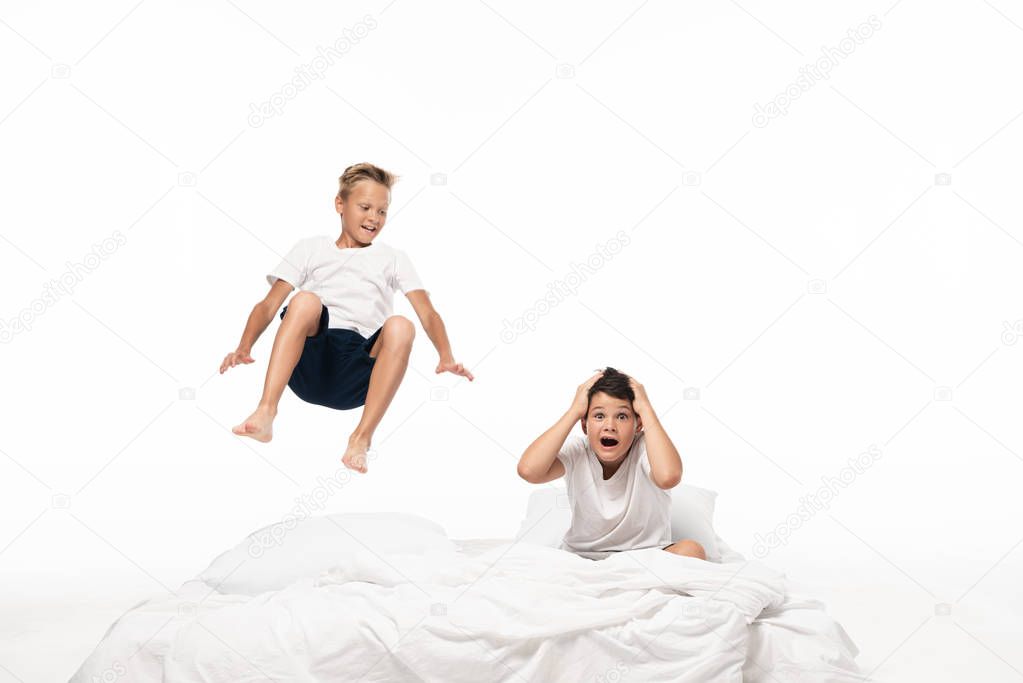 cheerful boy levitating over shocked brother sitting on bedding and holding hands on head isolated on white