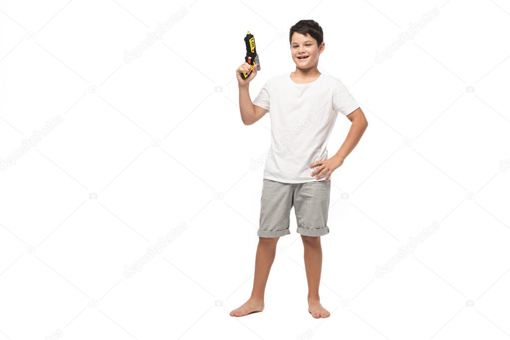 cheerful boy holding toy gun while standing with hands on hips and smiling at camera on white background