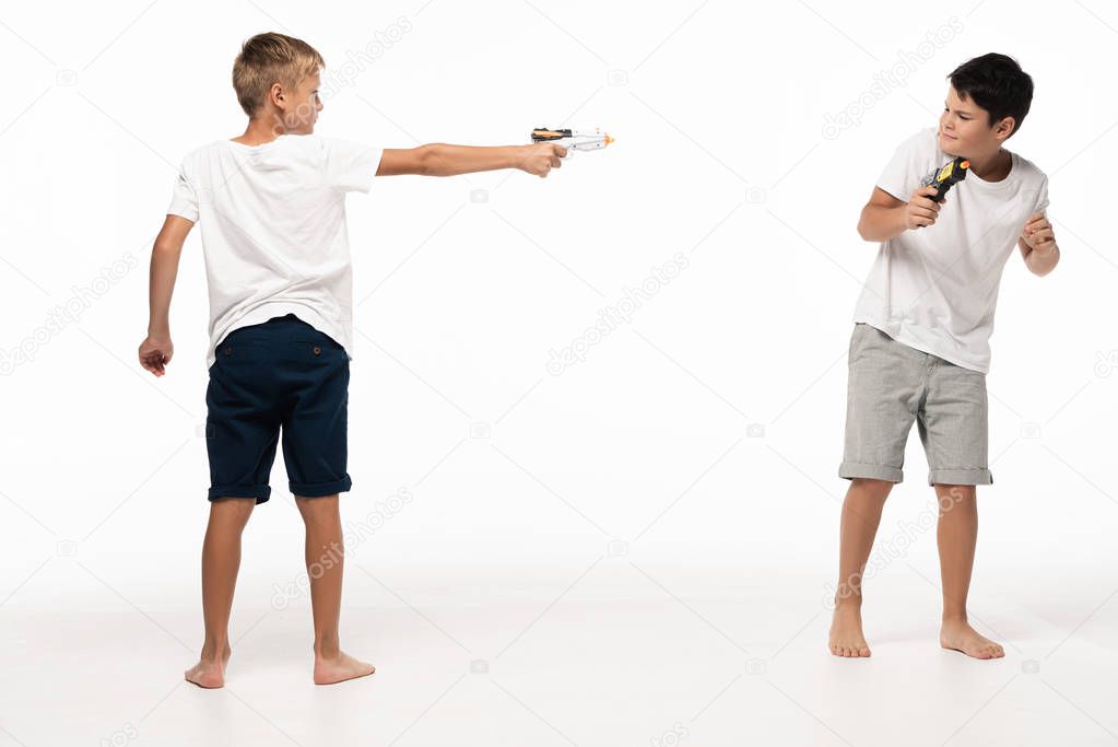  boy aiming with toy gun at scared brother on white background
