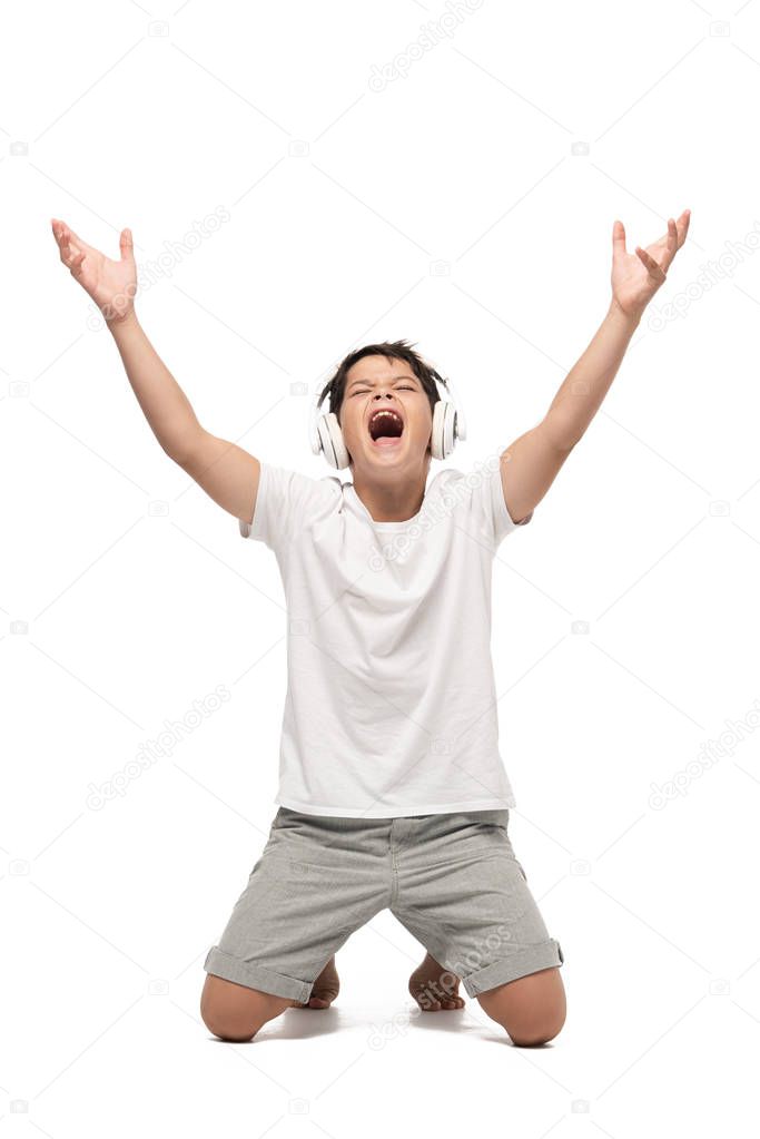 excited boy standing on knees with raised hands while listening music in headphones and singing on white background