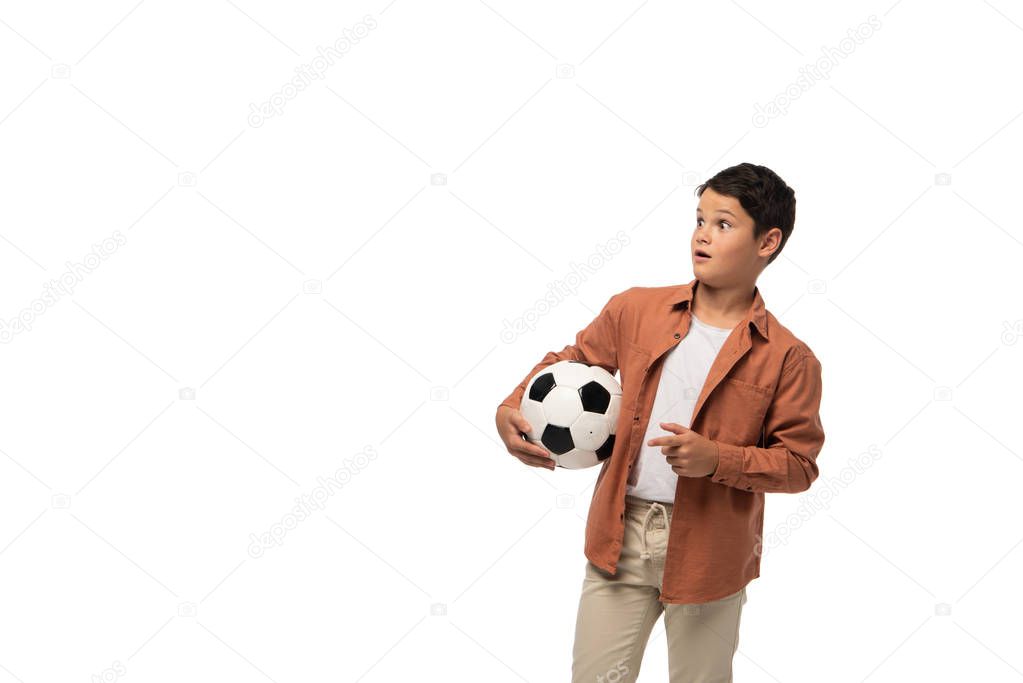 shocked boy holding soccer ball, looking away and pointing with finger isolated on white