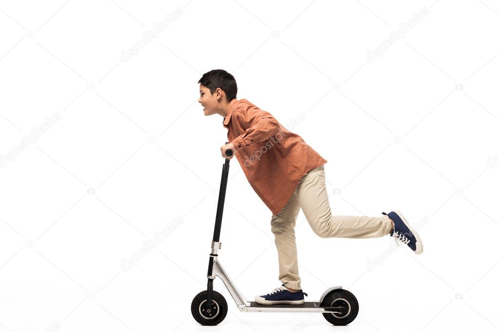 side view of smiling boy riding kick scooter on white background