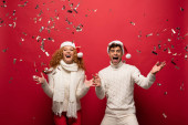 vzrušený pár v Santa klobouky křičí a slaví se zlatými konfety, izolované na červené