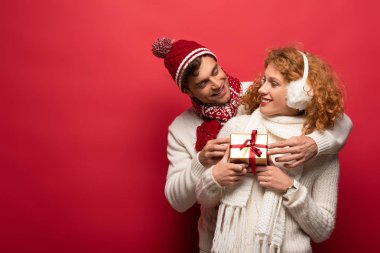 Kış kıyafetli mutlu çift, kırmızı ışıkta izole edilmiş bir Noel hediyesi taşıyor.