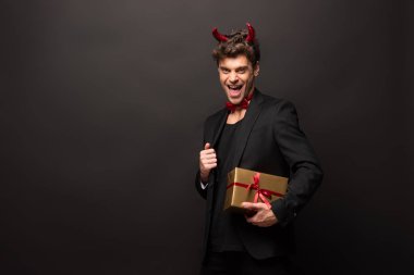 handsome excited man in devil costume holding gift boxes for halloween on black clipart