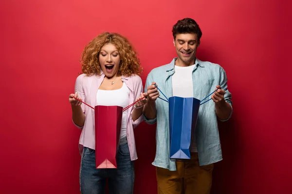 Couple Excité Regardant Dans Des Sacs Provisions Isolé Sur Rouge — Photo