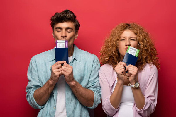 пара счастливых путешественников целующих паспорта с авиабилетами, изолированные на красный
