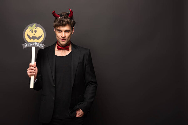 handsome man in devil costume holding pumpkin trick or treat sign on black