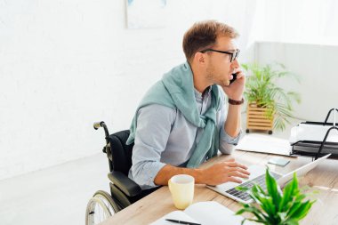 Tekerlekli sandalyedeki sıradan iş adamı akıllı telefonla konuşuyor ve masadaki bilgisayarı kullanıyor.