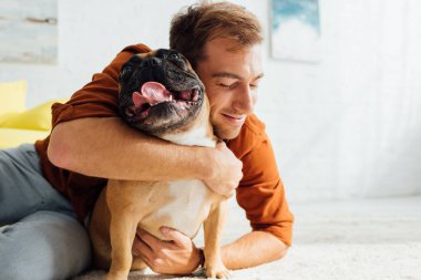 Gülen adam oturma odasında komik Fransız bulldog 'una sarılıyor.