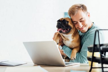 Gülümseyen adam dizüstü bilgisayarla çalışırken Fransız bulldog 'una sarılıyor.